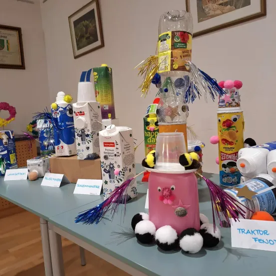 Ein Tisch ist gefüllt mit verschiedenen Gegenständen, darunter eine große pinke Spielzeugfigur mit einem weißen Hut, umgeben von bunten Kartons und Flaschen. Bilderrahmen schmücken die Wand.