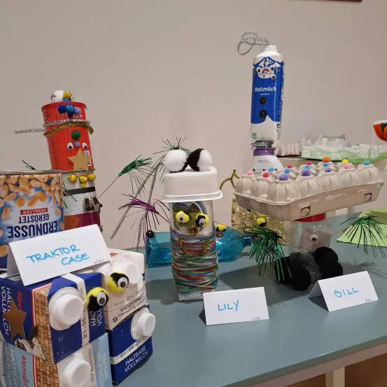 Auf einem Tisch sind verschiedene selbstgemachte Kreationen ausgestellt. Es gibt eine Schachtel mit Traktor-Design, eine weitere Schachtel mit Pinguin-Design und ein Glas mit bunten Fäden. Andere Gegenstände sind eine Flasche Vollmilch und ein Eierkarton. Jeder Gegenstand hat ein Namensschild.