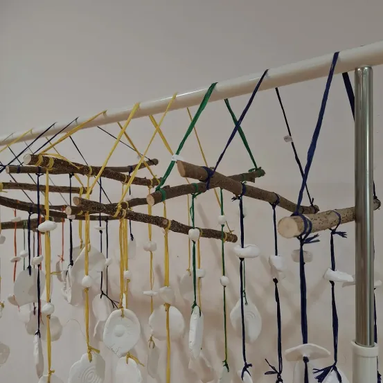 Eine Reihe von weißen Keramikobjekten hängt von verschiedenen farbigen Schnüren, die an einem weißen Hintergrund befestigt sind. Die Stöcke sind mit einem vertikalen weißen Pfosten verbunden.