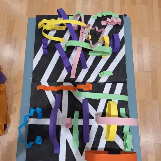 Ein farbenfrohes Papierkunstprojekt mit Zickzack- und Kreismustern ist auf einem Holzboden ausgestellt. Das Kunstwerk besteht aus verschiedenen farbigen Papieren, darunter lila, pink, gelb und grün.