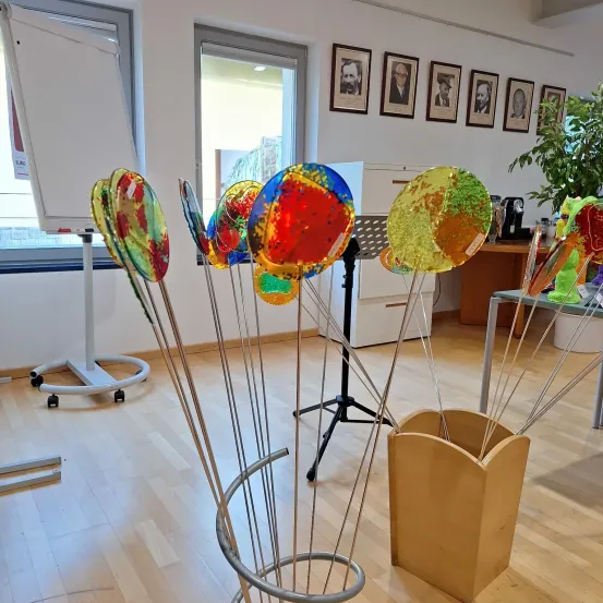 Ein Raum mit Holzboden, einer Glastür und einer Whiteboard. Bunte Glasobjekte auf Ständern. Gerahmte Bilder an der Wand.