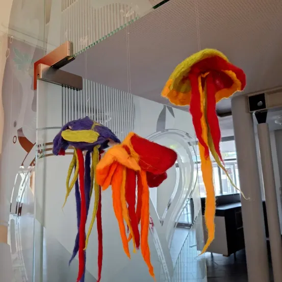 Zwei bunte Quallen-Dekorationen hängen von der Decke. Die linke ist lila und gelb. Die rechte ist gelb und rot. Sie befinden sich in einem Glasraum mit einem Spiraldesign.