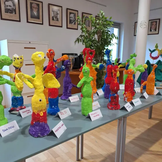 Ein Tisch zeigt verschiedene bunte Skulpturen mit Bezeichnungen wie Monster Lava, Shadow und anderen, vor dem Hintergrund von gerahmten Porträts und einer Pflanze.