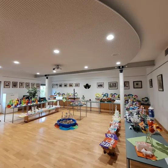 Ein Raum mit Holzböden hat mehrere Tische mit Kunstausstellungen aufgestellt. An der Wand hängen gerahmte Bilder und eine Hängelampe hängt in der Mitte.