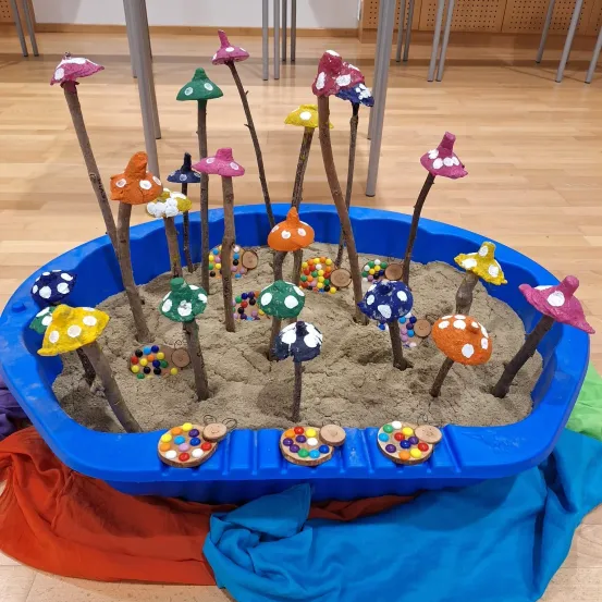Ein blaues Becken gefüllt mit Sand zeigt verschiedene farbenfrohe Pilzkunstwerke mit einigen verstreuten Süßigkeiten. Die Pilze haben unterschiedliche Farben und Designs, wobei einige Stiele wie Stöcke haben. Der Sand ist gleichmäßig im Becken verteilt.