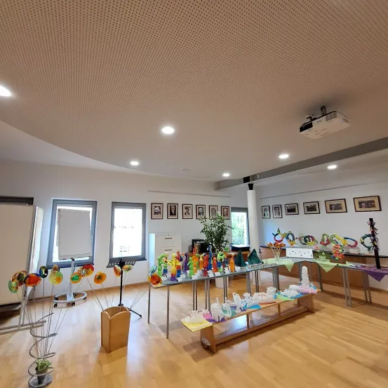 Ein leerer Raum mit Holzböden, weißen Wänden und einer Projektordecke. Präsentationstische mit bunten Kunstwerken, einer Tafel und gerahmten Bildern schmücken den Raum.