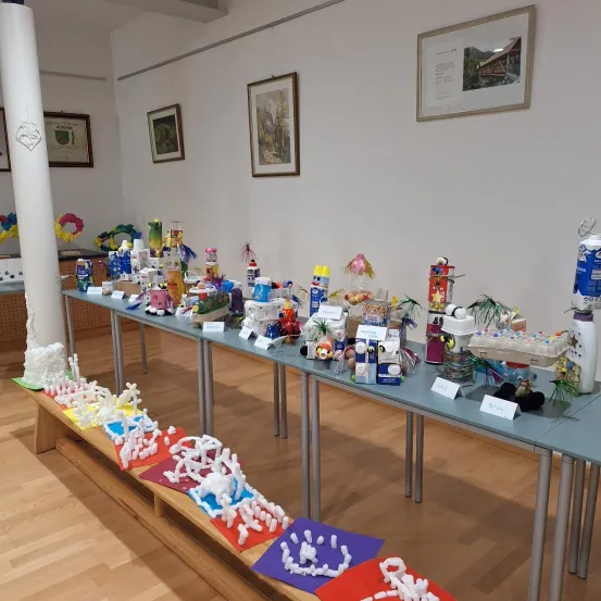 Ein Innenraum mit Holzböden, der zahlreiche bunte Skulpturen auf langen Tischen ausstellt, mit gerahmten Bildern an der Wand.