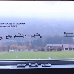 Ein Fensterausblick zeigt ein großes, baumbestandenes Feld mit einem Haus. Unter dem Fenster sind mehrere Papiere und ein Stift platziert. Ein Gebäudeplan ist über dem Feld zu sehen.