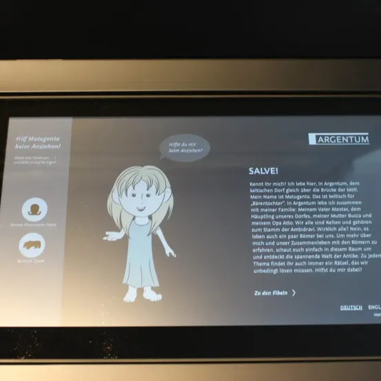 Ein Bildschirm zeigt ein Bild von einem Mädchen mit den Worten Salve und einer Hilfetaste. Darunter steht ein Text über ein Mädchen namens Matugenta, die Argentum liebt und ihre Familie. Der Bildschirm hat Optionen für Deutsch und Englisch.