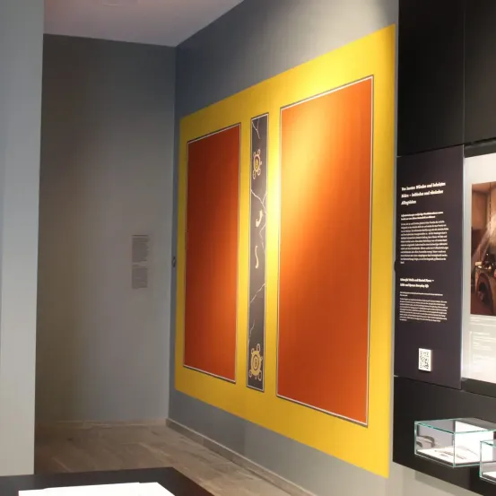 Eine Ausstellungswand zeigt drei große orangefarbene Tafeln mit gelben Rändern und abstrakten Mustern, begleitet von Texttafeln auf der rechten Seite. Der Raum hat einen Holzboden und eine Glasvitrine auf der rechten Seite.