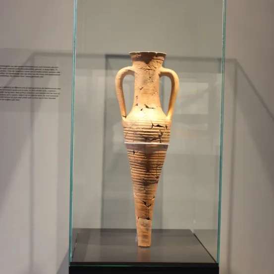 Eine alte, verwitterte Tonvase mit zwei Henkeln steht prominent in einer Glasvitrine, wahrscheinlich im Museum ausgestellt.