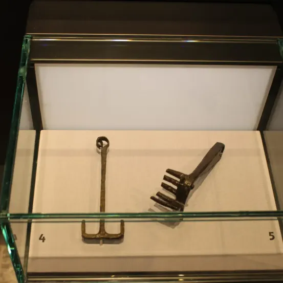 Ein Glasvitrine zeigt zwei antike Metallwerkzeuge, eine Harke und einen Dreizack, beschriftet mit den Nummern 4 und 5.
