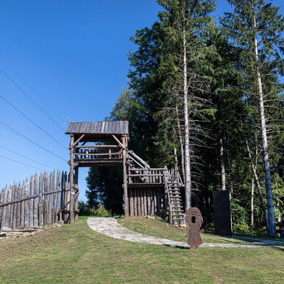 Ein hölzerner Aussichtsturm mit Treppen steht auf einem grasbewachsenen Hügel, umgeben von hohen Bäumen. Ein Steinweg führt zum Turm, mit einem Holzzaun und einer kleinen Statue in der Nähe.