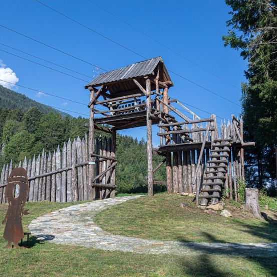 Eine Holzkonstruktion mit Treppen und Dach steht in einem Grasfeld mit Zaun. Dahinter befindet sich eine Bergkette.