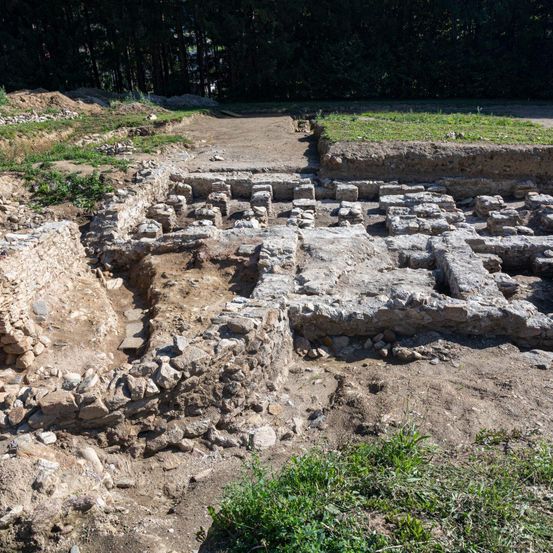 Eine archäologische Stätte mit verstreuten Steinen und Ruinen. Ein staubiger Pfad verläuft durch die Stätte, und an den Seiten ist Grün zu sehen.