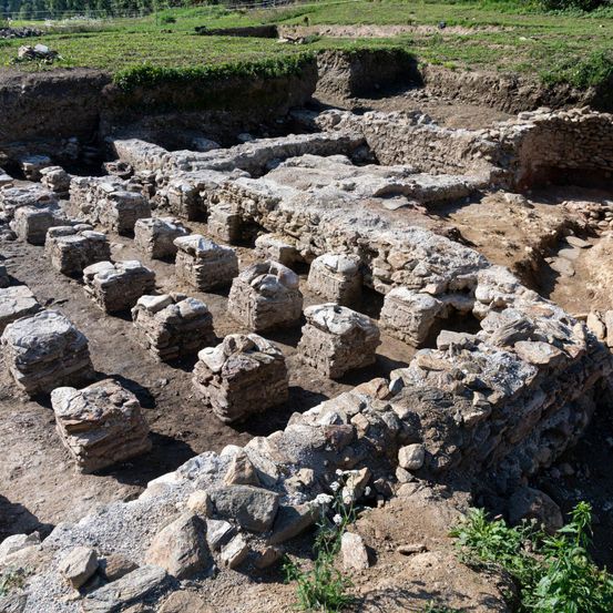 Eine archäologische Ausgrabungsstätte zeigt Überreste alter Steinstrukturen mit verstreuten Felsen und Erde. Im Hintergrund ist ein grünes Feld zu sehen.