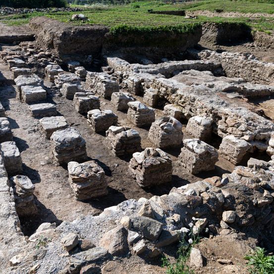 Eine archäologische Stätte mit angeordneten Steinblöcken und Erde. Einige Steine sind gestapelt, andere verstreut. Im Hintergrund sind grünes Gras und Pflanzen sichtbar.