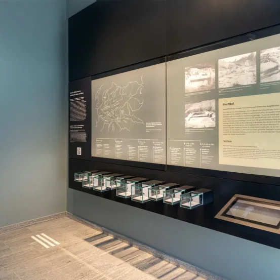 Ein Museumsaussteller mit einer Karte und Informationstafeln. Unter den Tafeln befinden sich Glasvitrinen mit verschiedenen Artefakten. Der Raum hat einen sauberen, modernen Look mit einem gefliesten Boden.