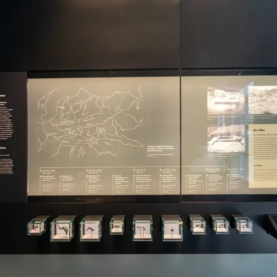 Eine Museumsausstellung zeigt eine Karte des alten Roms mit verschiedenen beschrifteten Orten. Daneben befindet sich ein Bild eines alten Gebäudes. Unter der Karte stehen mehrere Glasboxen mit kleinen Skulpturen darin.