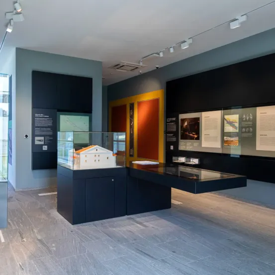 In einem Museum wird ein kleines Gebäudemodell in einer Glasvitrine ausgestellt. Um die Vitrine herum befinden sich mehrere Informationstafeln an den Wänden. Der Raum hat ein modernes Design mit eleganten Schwarz- und Grautönen.