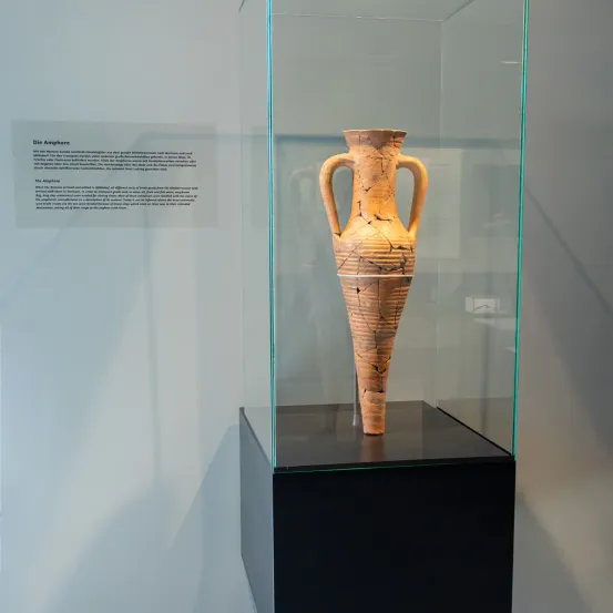 Eine antike Amphora aus dem Mittelalter in einer Glasvitrine, wahrscheinlich aus dem Mittelalter, wird in einem Museum ausgestellt. Sie hat zwei Henkel und zeigt Alters- und Abnutzungserscheinungen.