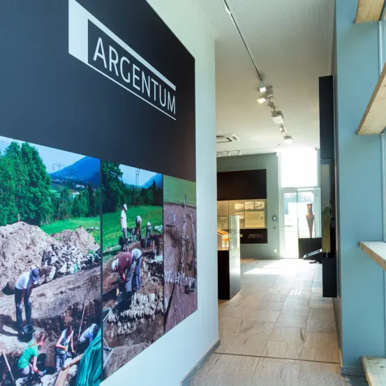 Eine Wand mit dem Wort Argentum in weißen Buchstaben. Im Hintergrund sind Fotos von Menschen beim Graben zu sehen. Der Boden ist aus Holz.