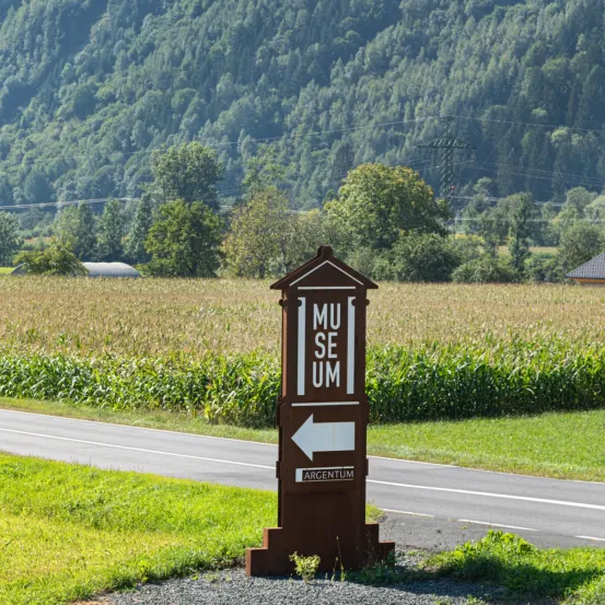 Ein Wegweiser am Straßenrand weist Besucher zum Museum Argentum. Der Schild befindet sich vor einer weitläufigen Feld- und Bergkulisse.