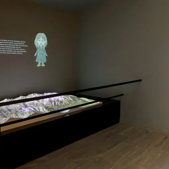 Eine Ausstellung mit einer Illustration eines Mädchens und einem 3D-Geländemodell auf einer Glasfläche, mit einem projizierten Bild von deutschem Text an einer Wand.