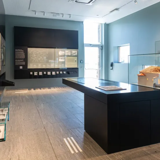 Ein Innenbereich mit einer schwarzen Ausstellungstafel, einer großen Karte und einem Glasschrank mit einem Modellhaus. Der Raum hat Holzböden und Wände, mit einer Tür, die nach draußen führt.