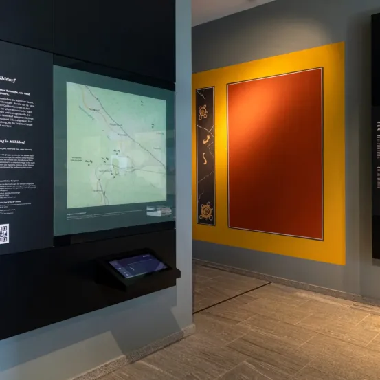 Eine Ausstellungsanzeige mit einem großen Bildschirm, der eine Karte und einen gelben und orangefarbenen Hintergrund zeigt. Die Karte zeigt ein ländliches Gebiet mit geografischen Merkmalen.