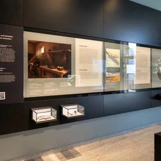 Eine Museumsausstellung zeigt ein Bild eines alten Raums mit Tisch und Fenster, neben informativen Texten und Illustrationen an den Wänden.