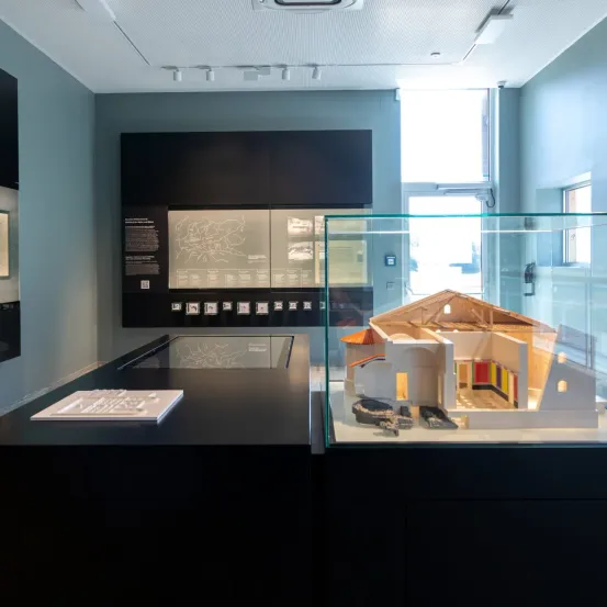 Ein Raum mit einer Glasvitrine, die ein Modell eines Gebäudes enthält. Das Modell befindet sich in einer Glasvitrine. Ein Buch liegt auf einem Tisch. Der Raum hat graue Wände.