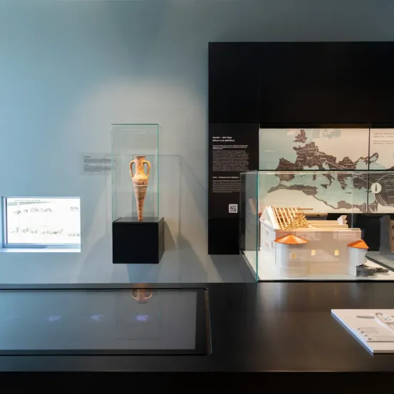 Eine Museumsausstellung zeigt eine antike Vase in einem Glaskasten und ein Modell eines Gebäudes in einem anderen Glaskasten. Im Hintergrund befinden sich eine Karte und Informationstafeln.