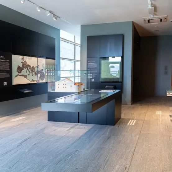 Ein Innenausstellungsbereich mit einem Glastisch, der eine Karte und ein kleines Gebäudemodell unter Glas zeigt. Die Wände sind dunkelblau und der Boden ist aus Holz.