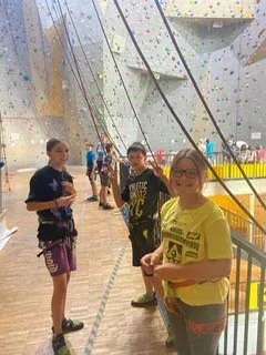 Vier Personen stehen auf einer Holzplattform in einem Kletterzentrum. Drei von ihnen tragen Gurte und lächeln. Die Wand hinter ihnen ist mit farbenfrohen Klettergriffen gefüllt.