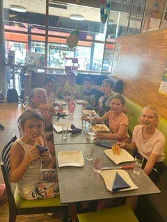 Eine Gruppe von Menschen sitzt an einem langen Tisch in einem Restaurant. Sie essen Pizza. Der Tisch hat Teller, Gläser und Servietten. Das Restaurant hat Holzwände und ein Glasfenster.