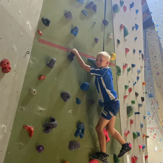 Ein junger Junge in einem blauen und weißen Shirt und Shorts klettert an einer Felswand mit vielen bunten Griffen.