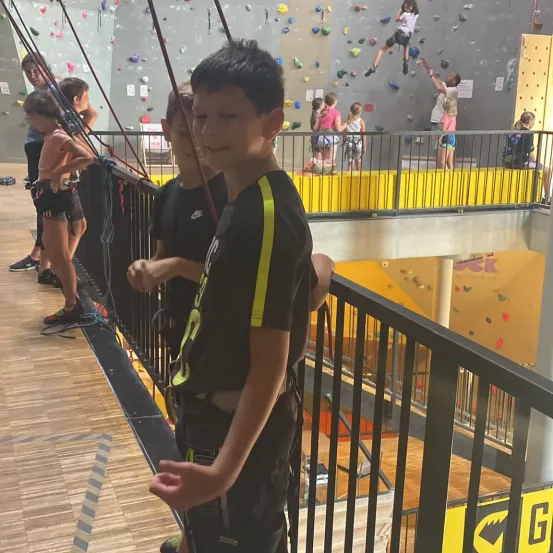 Kinder, die Gurte tragen, stehen auf einem Balkon und beobachten Kletterer an einer Kletterwand. Einige Kinder klettern, während andere zuschauen. Der Balkon hat schwarze Geländer.