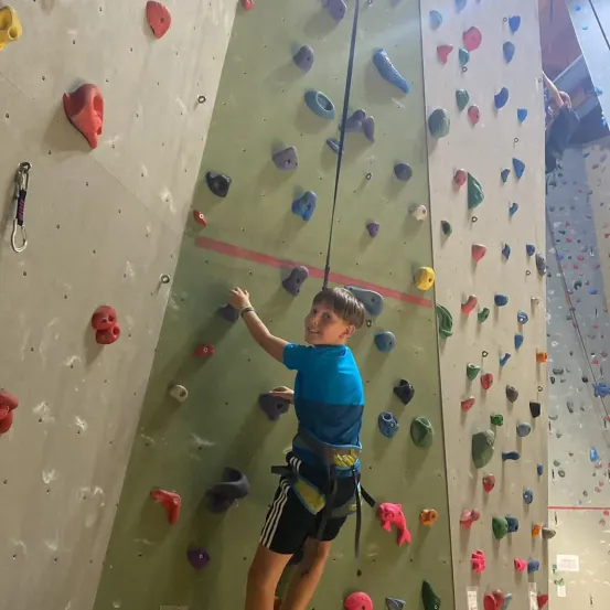 Ein junger Junge klettert an einer Kletterwand mit bunten Griffen, trägt ein blaues T-Shirt und schwarze Shorts. Er benutzt beide Hände und Füße, um die Griffe zu packen und aufzusteigen.