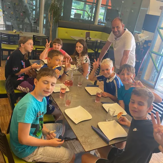 Eine Gruppe von Kindern und ein Mann sitzen an einem Tisch in einem Restaurant. Sie machen Friedenszeichen und lächeln.