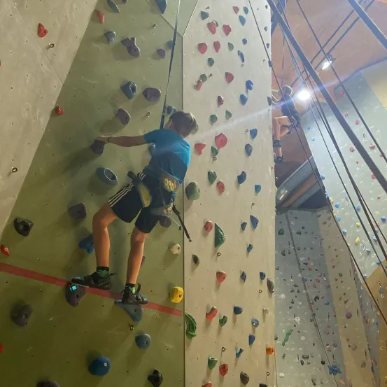 Ein junger Junge klettert an einer mit bunten Griffen bedeckten Wand, die mit einem Gurtzeug befestigt ist. Ein weiterer Kletterer ist hinter ihm, und die Wand ist von Lichtern beleuchtet.