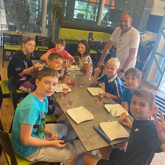 Eine Gruppe von Kindern und einem Mann sitzen um einen Tisch in einem Restaurant. Sie haben Teller, Gläser und Servietten auf dem Tisch. Sie scheinen ihre Mahlzeit zu genießen.