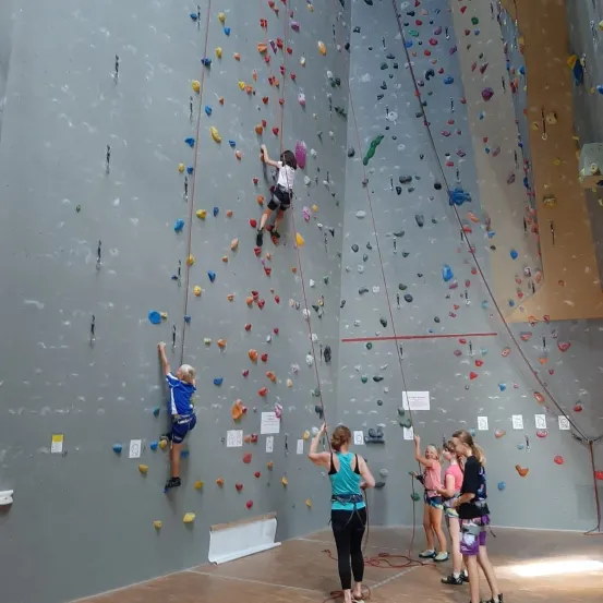 Mehrere Kinder klettern an einer Wand, gesichert durch Seile. Eine Frau beobachtet sie, während andere in der Nähe warten.