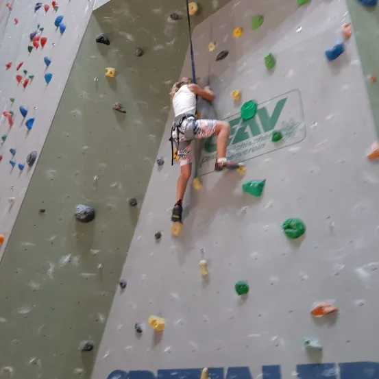 Eine Person klettert an einer Felswand mit verschiedenen farbigen Griffen hoch, trägt einen Gurt und Turnschuhe. Die Wand hat einen weißen Hintergrund mit unterschiedlich farbigen Griffen.