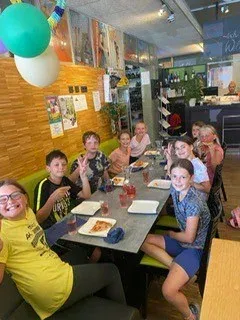 Eine Gruppe von Kindern sitzt an einem Tisch in einem Restaurant, lächelt und posiert für ein Foto. Es gibt Luftballons und Dekorationen an der Wand und Teller, Gläser und Essen auf dem Tisch.