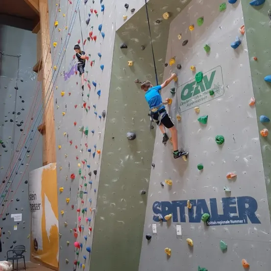 Zwei Kinder klettern an einer Kletterwand. Die Wand hat bunte Steine und das Wort "SPITALER" darauf geschrieben. Ein weiteres Wort ist in Blau geschrieben. Ein paar Leute stehen herum.