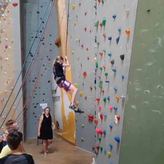 Ein junges Mädchen klettert an einer Felswand mit mehrfarbigen Griffen. Eine Frau in einem schwarzen Kleid beobachtet sie, während ein Junge und eine Frau in Gelb in der Nähe beobachten. Die Wand ist mit verschiedenen farbigen Griffen verziert.