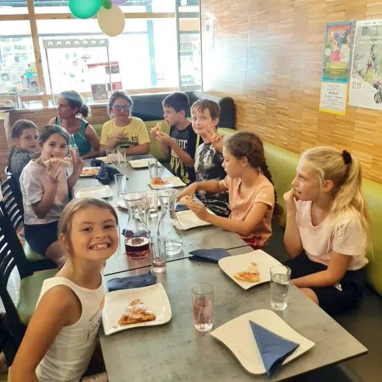 Eine Gruppe von Kindern sitzt am Tisch und isst Pizza, lächelt und schaut in die Kamera. Der Tisch ist mit Tellern, Gläsern und Servietten gedeckt. Dahinter hat die Wand Poster und Luftballons.