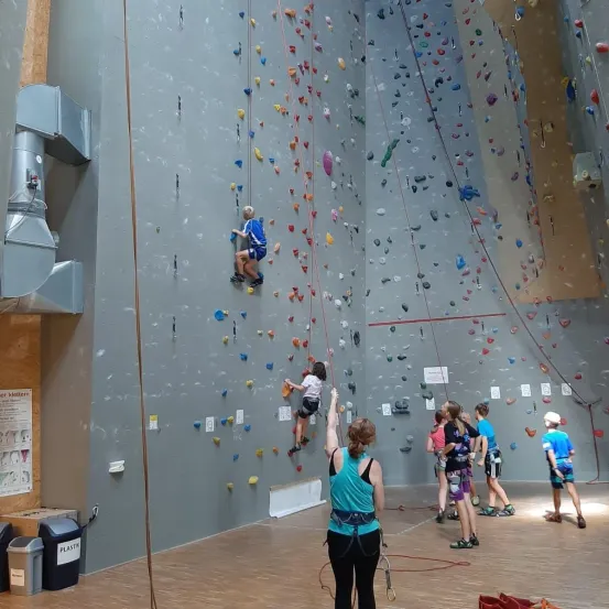 Indoor-Kletterwand, Kletterer besteigen die Wand, eine Frau unterstützt einen Kletterer, andere beobachten und stehen herum, bunte Griffe an der Wand.