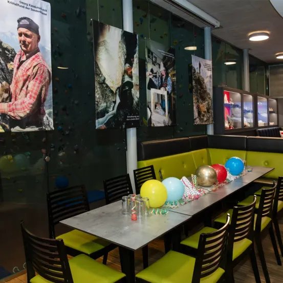 Ein Indoor-Kletterzentrum mit einem für eine Party gedeckten Tisch, gelben Stühlen, einem Metalltisch und bunten Luftballons und Dekorationen. Plakate von Bergsteigern hängen an der Wand.